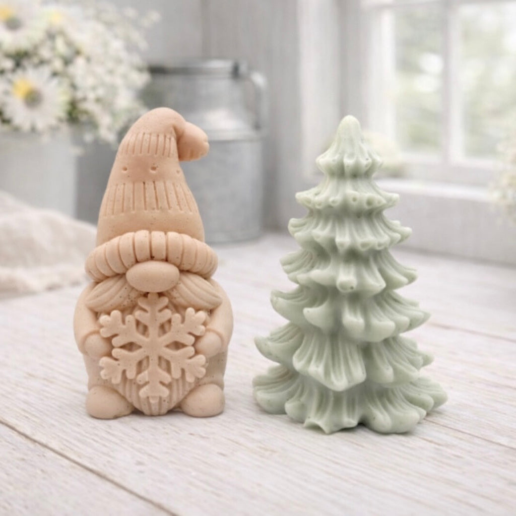 A pair of handmade vegan soaps featuring a Christmas gonk (gnome) with a textured hat holding a large snowflake, standing next to a pale mint-green 3D Christmas tree with layered, snow-dusted branches, displayed in a bright farmhouse setting.