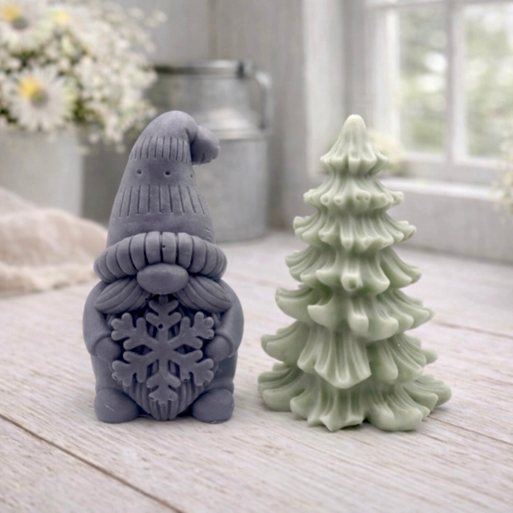 A pair of handmade vegan soaps featuring a Christmas gonk (gnome) with a textured hat holding a large snowflake, standing next to a pale mint-green 3D Christmas tree with layered, snow-dusted branches, displayed in a bright farmhouse setting.