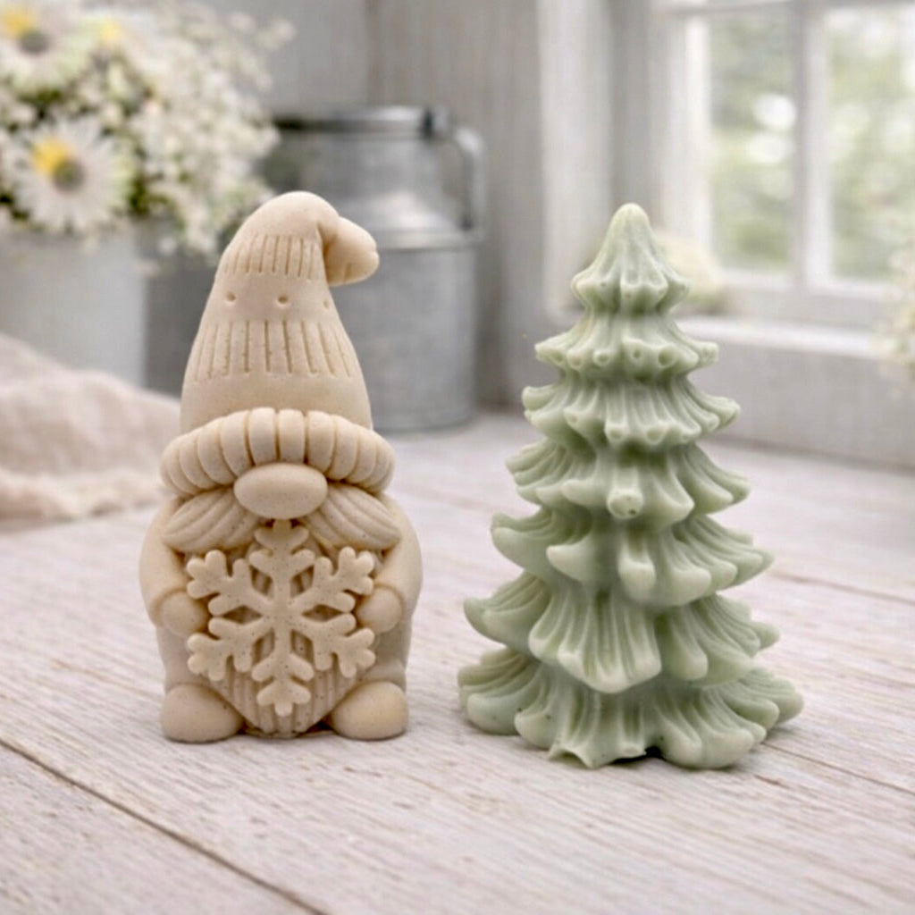 A pair of handmade vegan soaps featuring a Christmas gonk (gnome) with a textured hat holding a large snowflake, standing next to a pale mint-green 3D Christmas tree with layered, snow-dusted branches, displayed in a bright farmhouse setting.