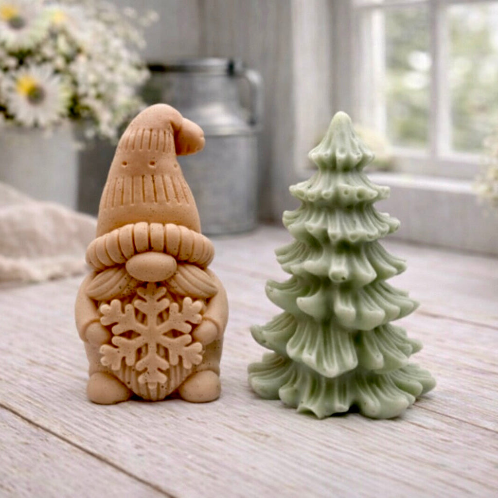A pair of handmade vegan soaps featuring a Christmas gonk (gnome) with a textured hat holding a large snowflake, standing next to a pale mint-green 3D Christmas tree with layered, snow-dusted branches, displayed in a bright farmhouse setting.