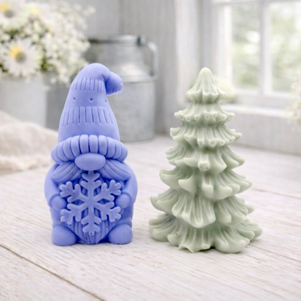 A pair of handmade vegan soaps featuring a Christmas gonk (gnome) with a textured hat holding a large snowflake, standing next to a pale mint-green 3D Christmas tree with layered, snow-dusted branches, displayed in a bright farmhouse setting.