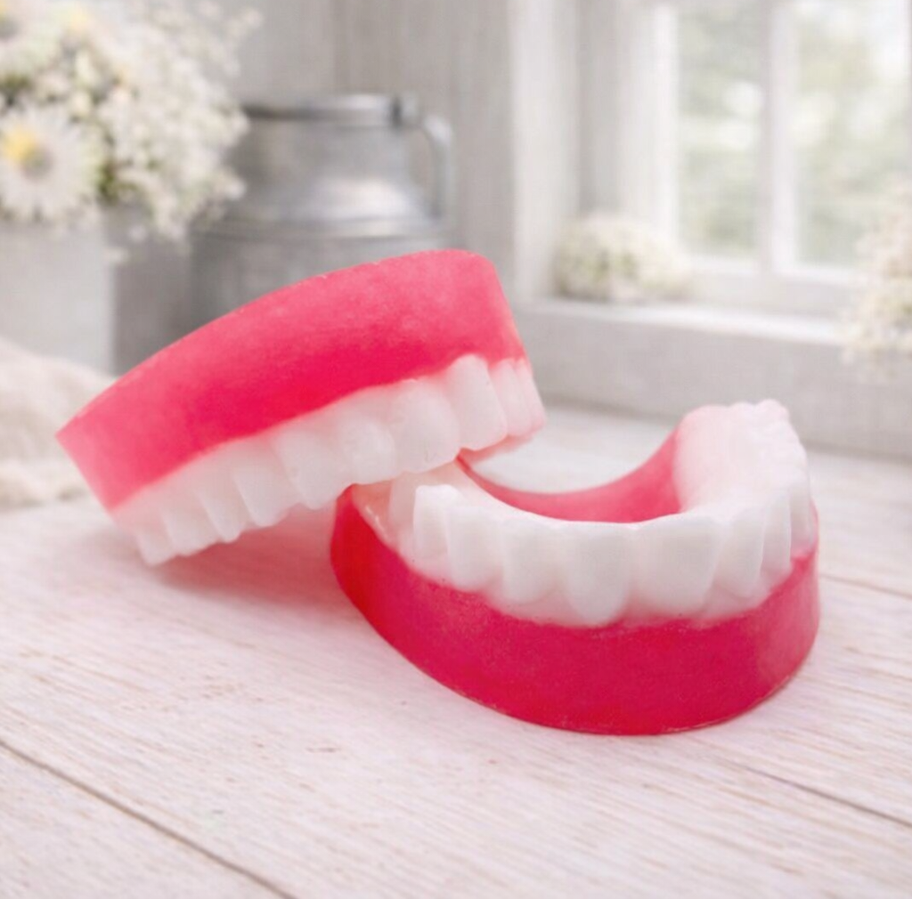 A 3D handmade vegan soap sculpted to look like a realistic set of dentures, featuring vibrant pink gums and a full row of white teeth, resting on a light wooden surface in a bright farmhouse setting.