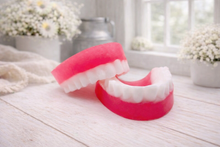 Load image into Gallery viewer, A 3D handmade vegan soap sculpted to look like a realistic set of dentures, featuring vibrant pink gums and a full row of white teeth, resting on a light wooden surface in a bright farmhouse setting.
