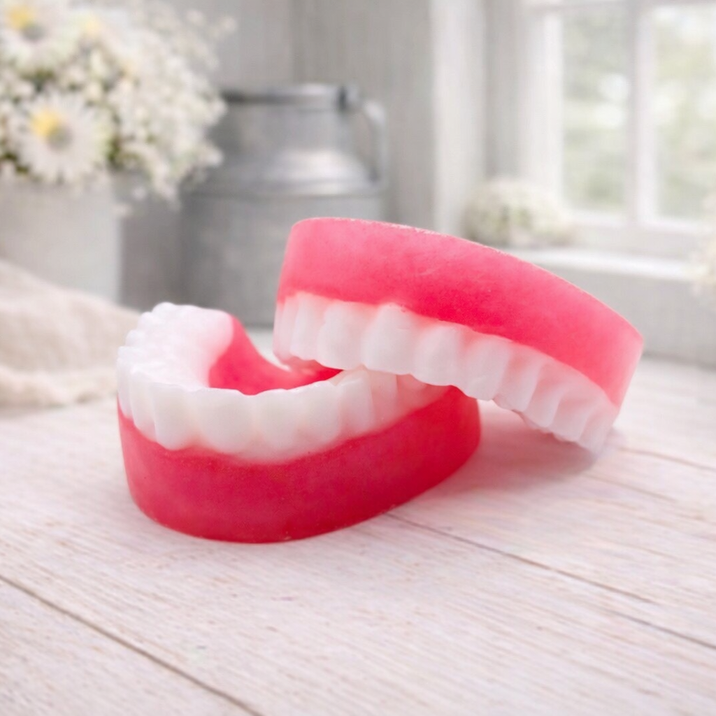 A 3D handmade vegan soap sculpted to look like a realistic set of dentures, featuring vibrant pink gums and a full row of white teeth, resting on a light wooden surface in a bright farmhouse setting.