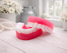 Load image into Gallery viewer, A 3D handmade vegan soap sculpted to look like a realistic set of dentures, featuring vibrant pink gums and a full row of white teeth, resting on a light wooden surface in a bright farmhouse setting.
