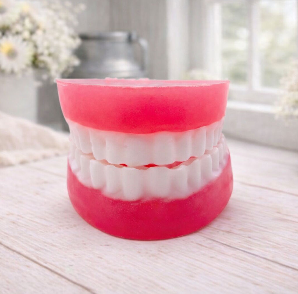 A 3D handmade vegan soap sculpted to look like a realistic set of dentures, featuring vibrant pink gums and a full row of white teeth, resting on a light wooden surface in a bright farmhouse setting.