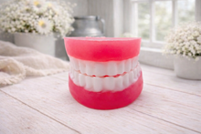 Load image into Gallery viewer, A 3D handmade vegan soap sculpted to look like a realistic set of dentures, featuring vibrant pink gums and a full row of white teeth, resting on a light wooden surface in a bright farmhouse setting.
