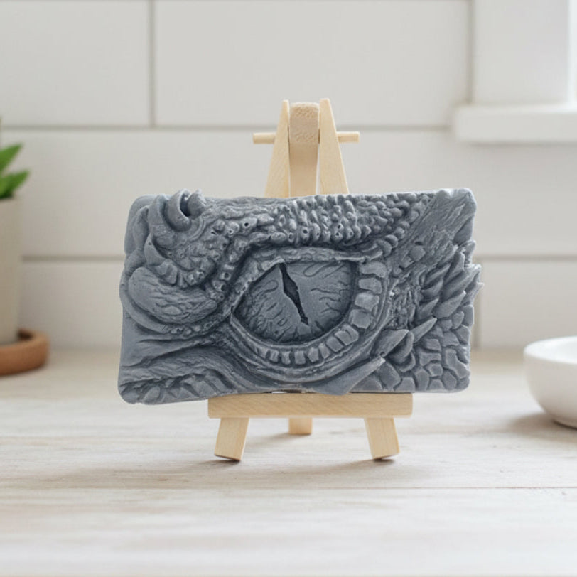 A rectangular handmade vegan soap in a slate-grey tone, featuring a highly detailed, close-up relief of a mythical dragon or dinosaur eye surrounded by thick, pebbled scales and reptilian skin textures, displayed on a miniature wooden easel.