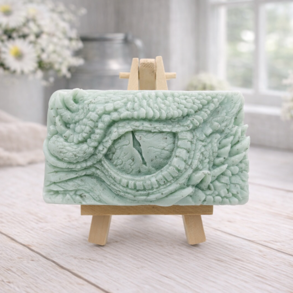 A rectangular handmade vegan soap in a slate-grey tone, featuring a highly detailed, close-up relief of a mythical dragon or dinosaur eye surrounded by thick, pebbled scales and reptilian skin textures, displayed on a miniature wooden easel.