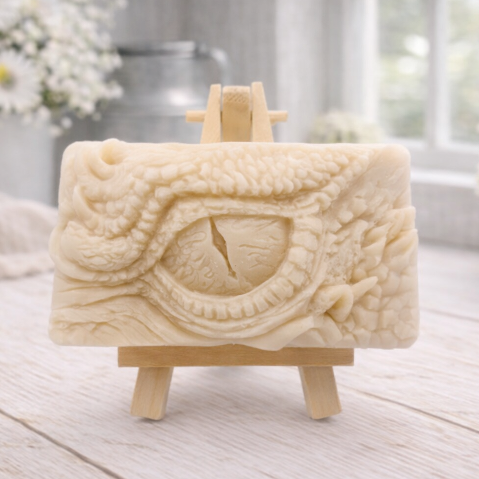A rectangular handmade vegan soap in a slate-grey tone, featuring a highly detailed, close-up relief of a mythical dragon or dinosaur eye surrounded by thick, pebbled scales and reptilian skin textures, displayed on a miniature wooden easel.