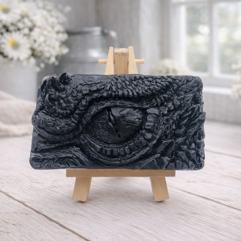 A rectangular handmade vegan soap in a slate-grey tone, featuring a highly detailed, close-up relief of a mythical dragon or dinosaur eye surrounded by thick, pebbled scales and reptilian skin textures, displayed on a miniature wooden easel.