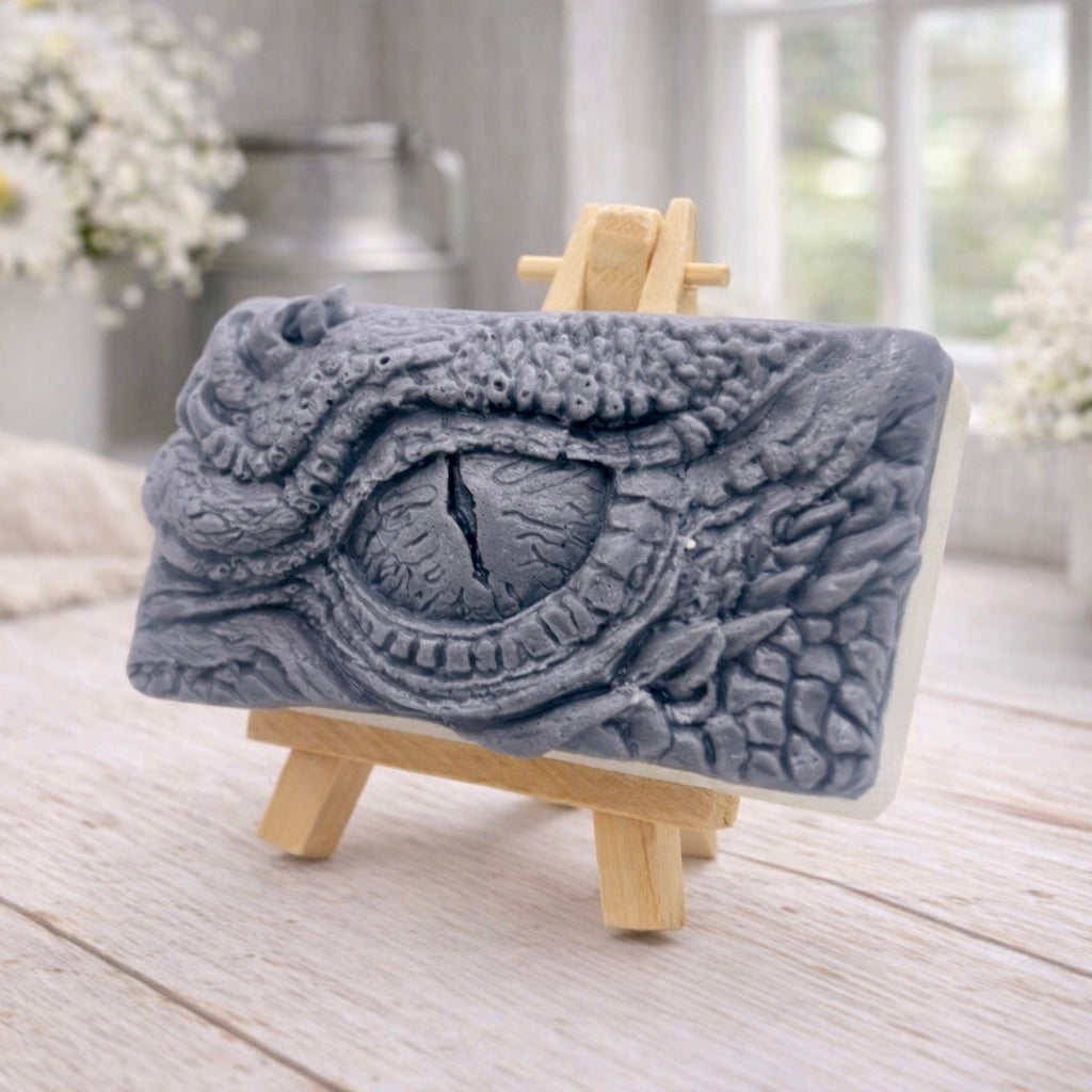 A rectangular handmade vegan soap in a slate-grey tone, featuring a highly detailed, close-up relief of a mythical dragon or dinosaur eye surrounded by thick, pebbled scales and reptilian skin textures, displayed on a miniature wooden easel.