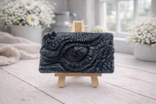 Load image into Gallery viewer, A rectangular handmade vegan soap in a slate-grey tone, featuring a highly detailed, close-up relief of a mythical dragon or dinosaur eye surrounded by thick, pebbled scales and reptilian skin textures, displayed on a miniature wooden easel.
