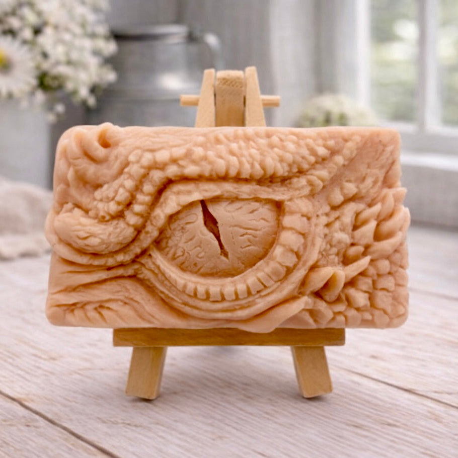 A rectangular handmade vegan soap in a slate-grey tone, featuring a highly detailed, close-up relief of a mythical dragon or dinosaur eye surrounded by thick, pebbled scales and reptilian skin textures, displayed on a miniature wooden easel.