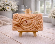 Load image into Gallery viewer, A rectangular handmade vegan soap in a slate-grey tone, featuring a highly detailed, close-up relief of a mythical dragon or dinosaur eye surrounded by thick, pebbled scales and reptilian skin textures, displayed on a miniature wooden easel.
