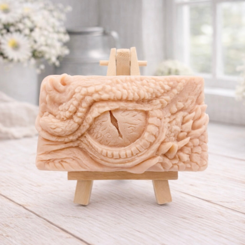 A rectangular handmade vegan soap in a slate-grey tone, featuring a highly detailed, close-up relief of a mythical dragon or dinosaur eye surrounded by thick, pebbled scales and reptilian skin textures, displayed on a miniature wooden easel.