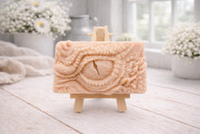 Load image into Gallery viewer, A rectangular handmade vegan soap in a slate-grey tone, featuring a highly detailed, close-up relief of a mythical dragon or dinosaur eye surrounded by thick, pebbled scales and reptilian skin textures, displayed on a miniature wooden easel.
