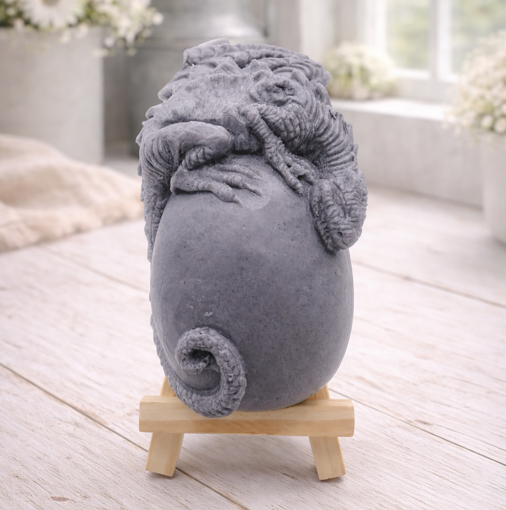 A 3D handmade vegan soap in a charcoal-grey tone, sculpted as a large prehistoric egg with a small dinosaur or dragon clinging to the top, featuring detailed scales and a curled tail, displayed on a miniature wooden easel.
