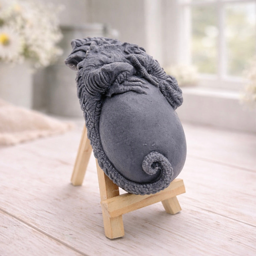 A 3D handmade vegan soap in a charcoal-grey tone, sculpted as a large prehistoric egg with a small dinosaur or dragon clinging to the top, featuring detailed scales and a curled tail, displayed on a miniature wooden easel.