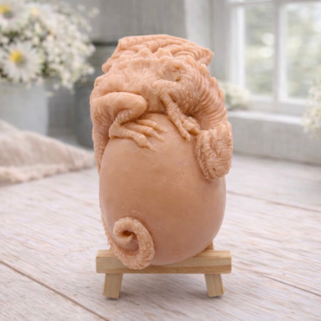 A 3D handmade vegan soap in a charcoal-grey tone, sculpted as a large prehistoric egg with a small dinosaur or dragon clinging to the top, featuring detailed scales and a curled tail, displayed on a miniature wooden easel.