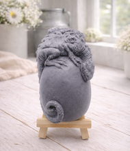 Load image into Gallery viewer, A 3D handmade vegan soap in a charcoal-grey tone, sculpted as a large prehistoric egg with a small dinosaur or dragon clinging to the top, featuring detailed scales and a curled tail, displayed on a miniature wooden easel.
