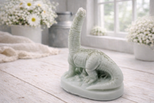 Load image into Gallery viewer, A handmade vegan soap in the shape of a long-necked brachiosaurus  dinosaur, featuring a soft mint green colour with natural speckles, set against a bright window and white flowers
