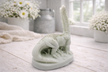 Load image into Gallery viewer, A handmade vegan soap in the shape of a long-necked brachiosaurus  dinosaur, featuring a soft mint green colour with natural speckles, set against a bright window and white flowers
