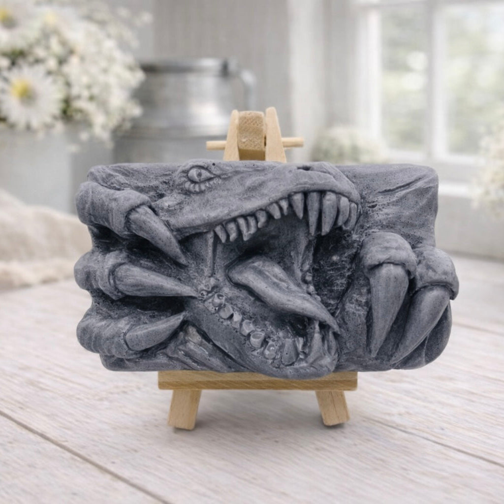 A rectangular handmade vegan soap in a charcoal-grey tone, featuring a high-relief carving of a Tyrannosaurus Rex with its jaws wide open in a roar and sharp claws framing the face, displayed on a miniature wooden easel.