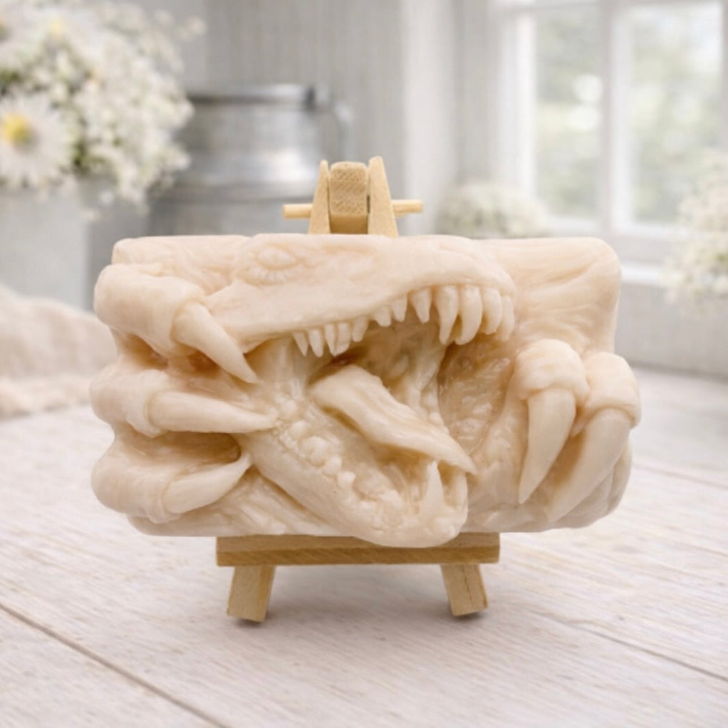 A rectangular handmade vegan soap  featuring a high-relief carving of a Tyrannosaurus Rex with its jaws wide open in a roar and sharp claws framing the face, displayed on a miniature wooden easel.