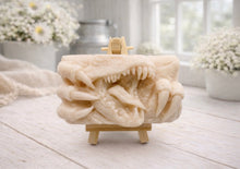 Load image into Gallery viewer, A rectangular handmade vegan soap  featuring a high-relief carving of a Tyrannosaurus Rex with its jaws wide open in a roar and sharp claws framing the face, displayed on a miniature wooden easel.
