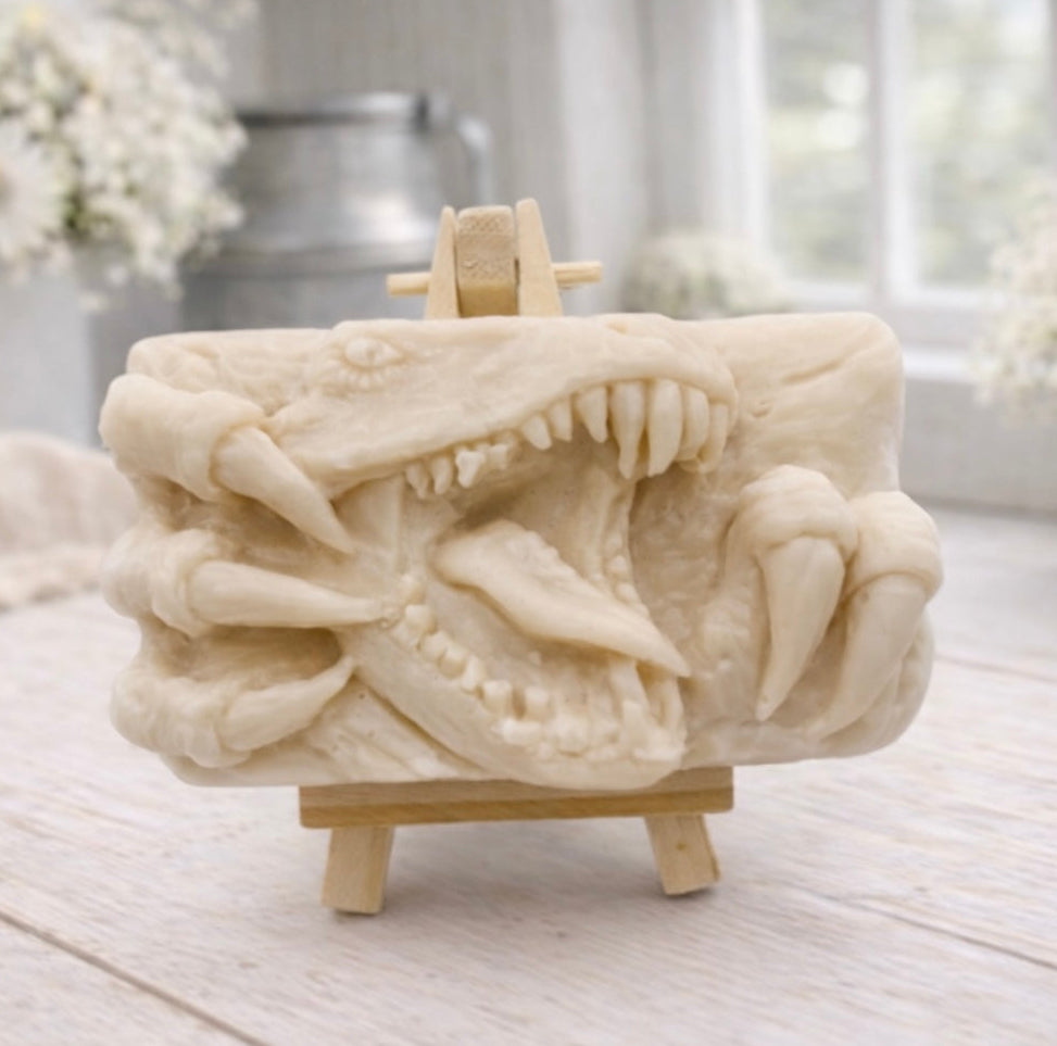 A rectangular handmade vegan soap  featuring a high-relief carving of a Tyrannosaurus Rex with its jaws wide open in a roar and sharp claws framing the face, displayed on a miniature wooden easel.