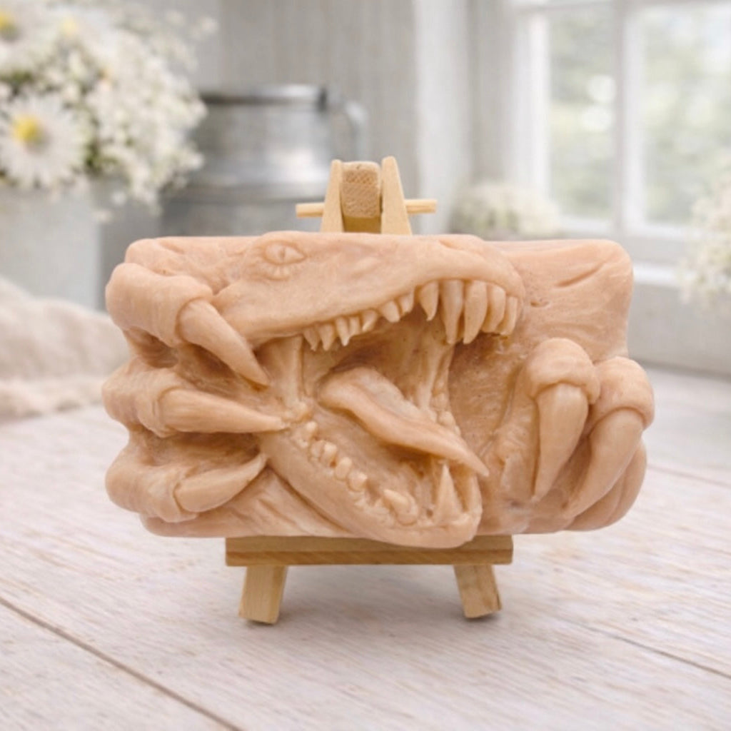 A rectangular handmade vegan soap  featuring a high-relief carving of a Tyrannosaurus Rex with its jaws wide open in a roar and sharp claws framing the face, displayed on a miniature wooden easel.