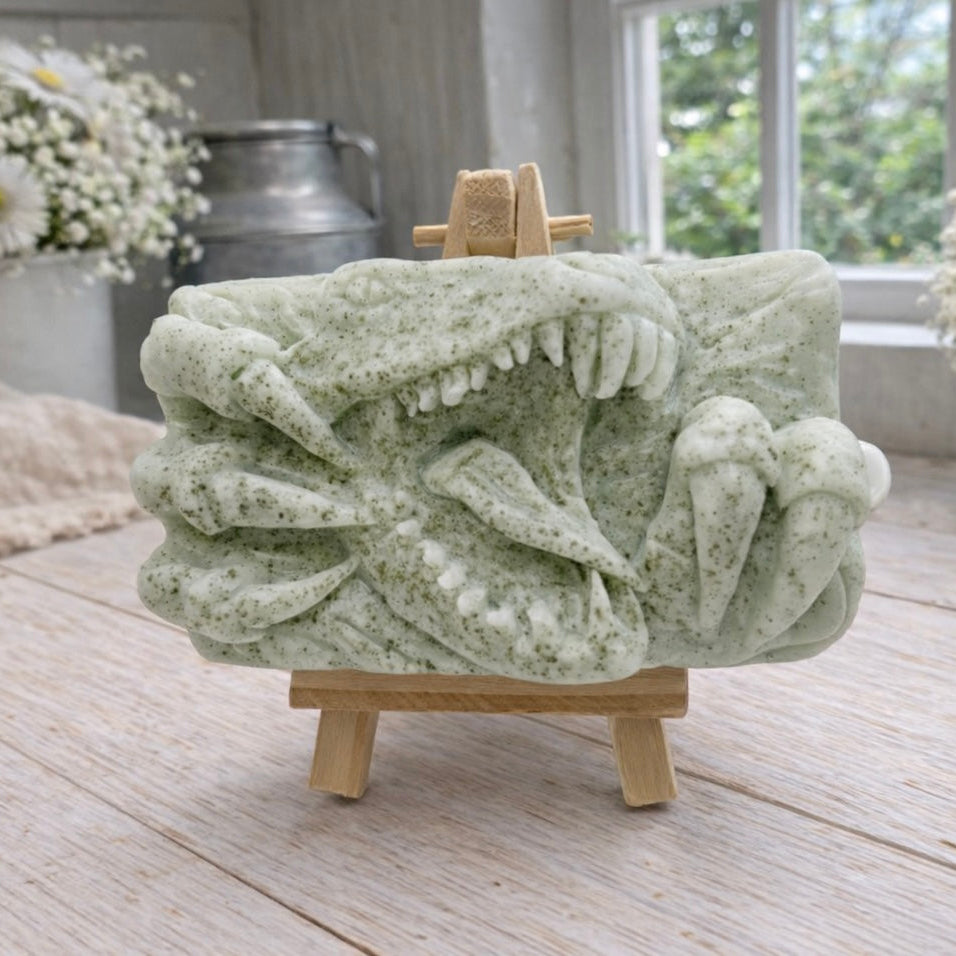 A rectangular handmade vegan soap  featuring a high-relief carving of a Tyrannosaurus Rex with its jaws wide open in a roar and sharp claws framing the face, displayed on a miniature wooden easel.