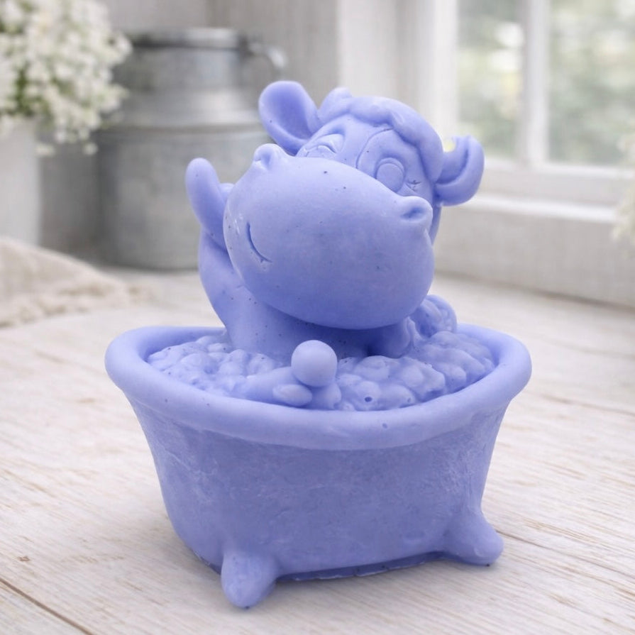 A handmade vegan soap is sculpted in the shape of a "dirty cow" character happily sitting in a bubble-filled roll-top bathtub with one hoof raised, displayed on a light wooden surface.