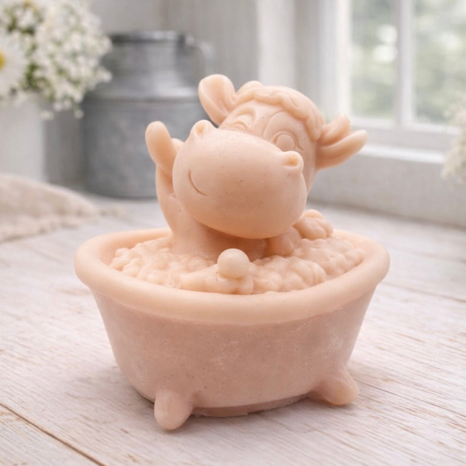 A handmade vegan soap is sculpted in the shape of a "dirty cow" character happily sitting in a bubble-filled roll-top bathtub with one hoof raised, displayed on a light wooden surface.
