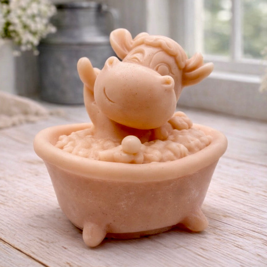 A handmade vegan soap is sculpted in the shape of a "dirty cow" character happily sitting in a bubble-filled roll-top bathtub with one hoof raised, displayed on a light wooden surface.
