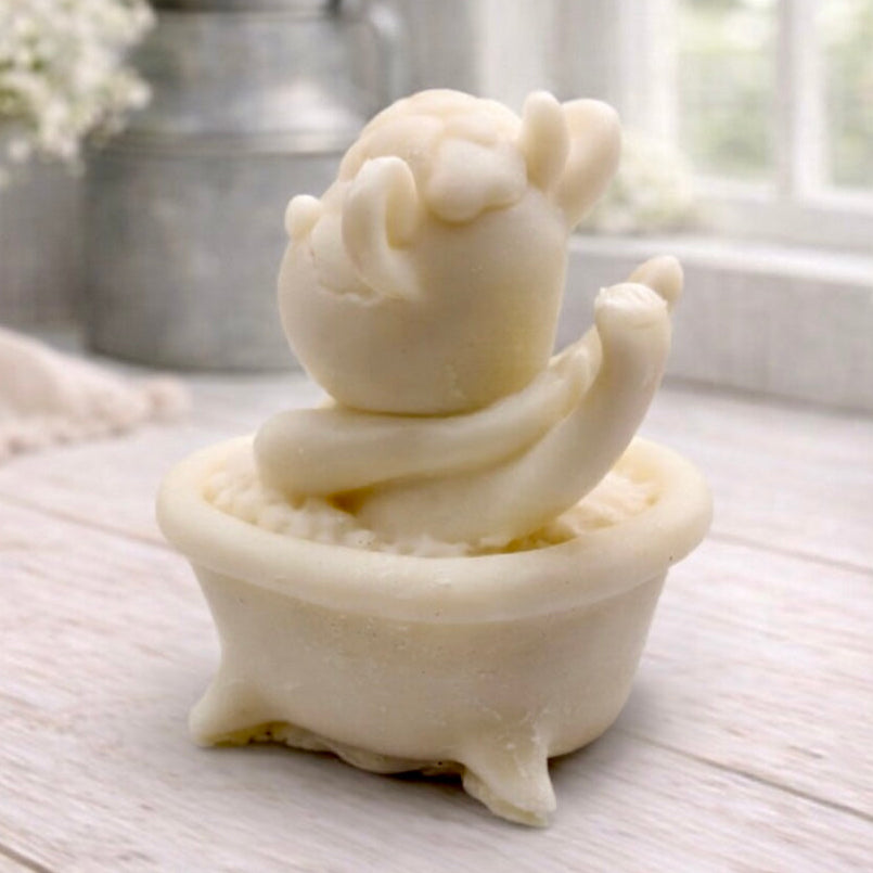 A handmade vegan soap is sculpted in the shape of a "dirty cow" character happily sitting in a bubble-filled roll-top bathtub with one hoof raised, displayed on a light wooden surface.