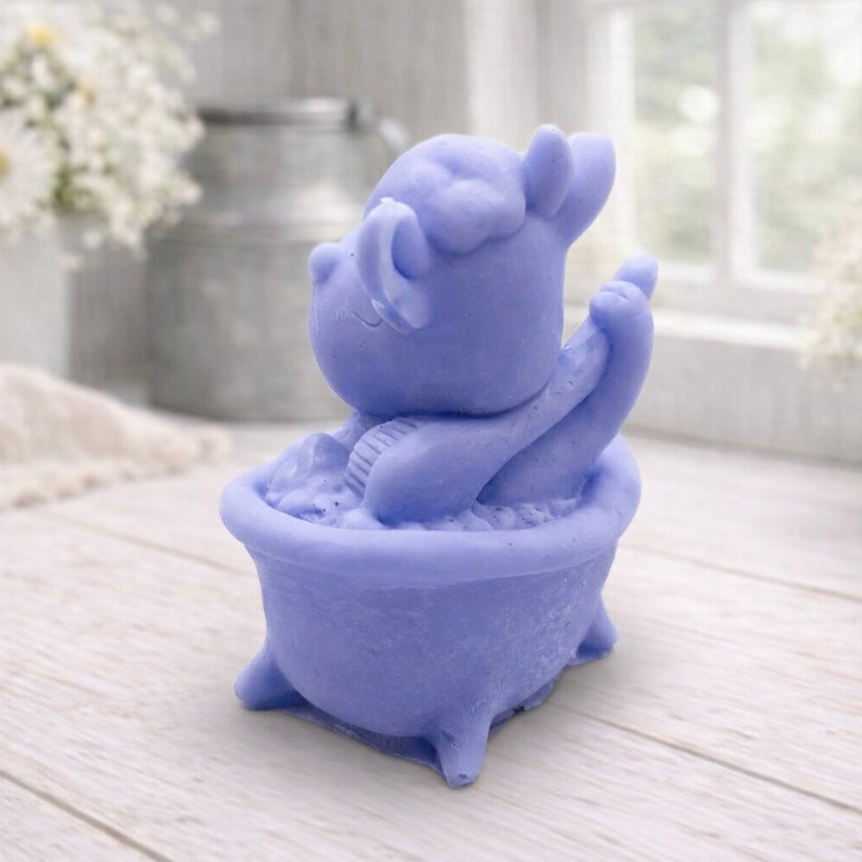 A handmade vegan soap is sculpted in the shape of a "dirty cow" character happily sitting in a bubble-filled roll-top bathtub with one hoof raised, displayed on a light wooden surface.