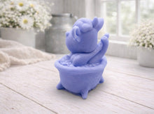 Load image into Gallery viewer, A handmade vegan soap is sculpted in the shape of a "dirty cow" character happily sitting in a bubble-filled roll-top bathtub with one hoof raised, displayed on a light wooden surface.
