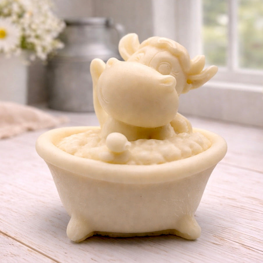 A handmade vegan soap is sculpted in the shape of a "dirty cow" character happily sitting in a bubble-filled roll-top bathtub with one hoof raised, displayed on a light wooden surface.