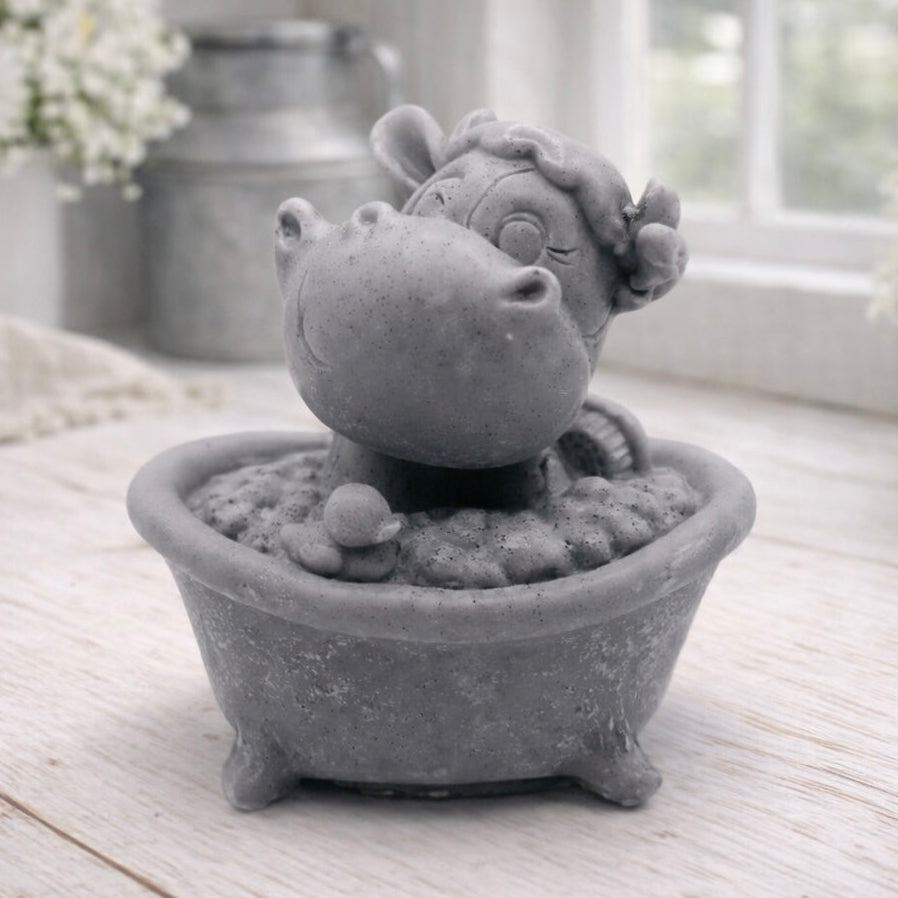 A handmade vegan soap is sculpted in the shape of a "dirty cow" character happily sitting in a bubble-filled roll-top bathtub with one hoof raised, displayed on a light wooden surface.