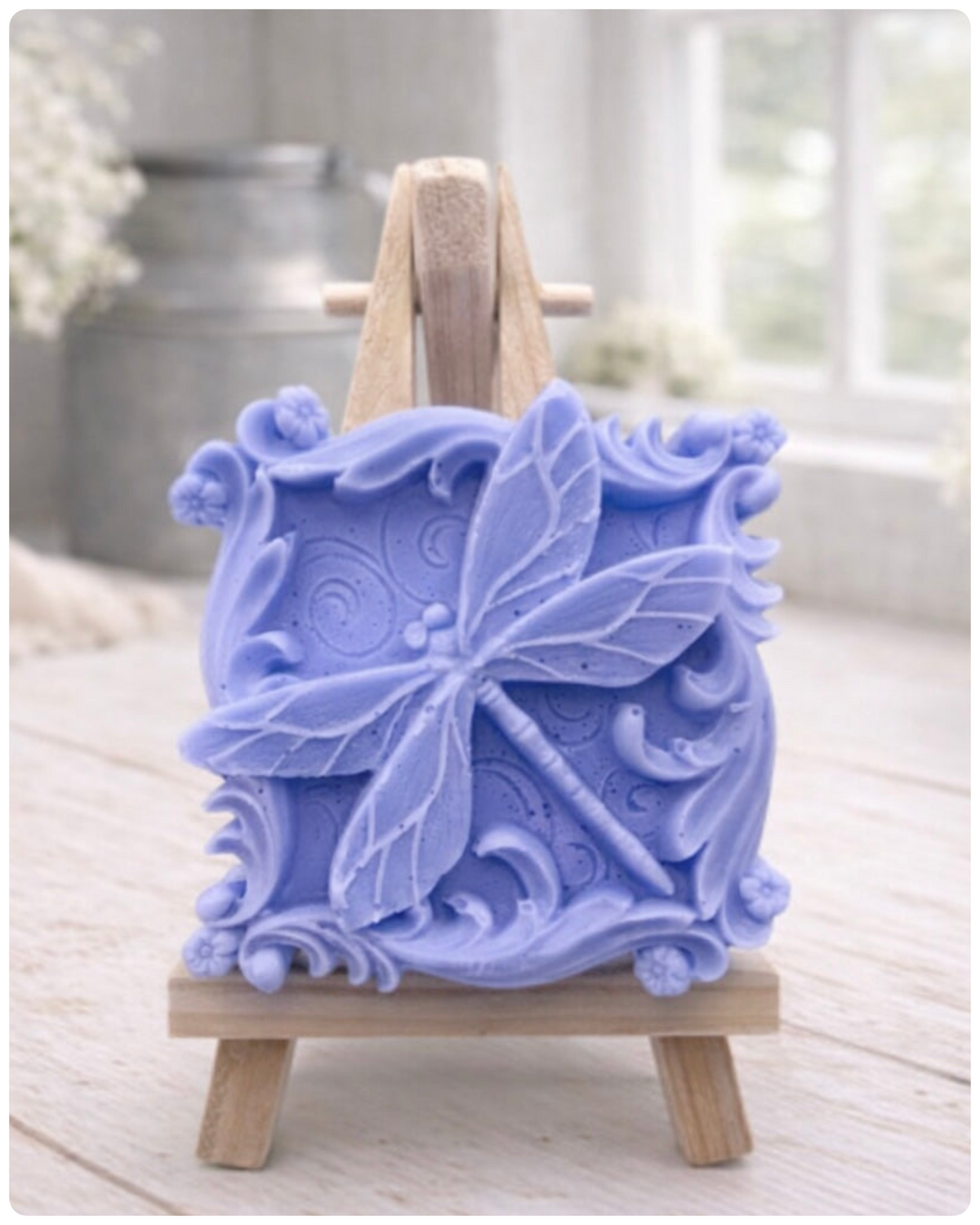 A beautiful handmade vegan soap in a natural tone, featuring a highly detailed relief of a dragonfly with delicate, outspread wings and a textured body, displayed on a miniature wooden easel against a soft farmhouse background.