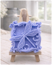 Load image into Gallery viewer, A beautiful handmade vegan soap in a natural tone, featuring a highly detailed relief of a dragonfly with delicate, outspread wings and a textured body, displayed on a miniature wooden easel against a soft farmhouse background.
