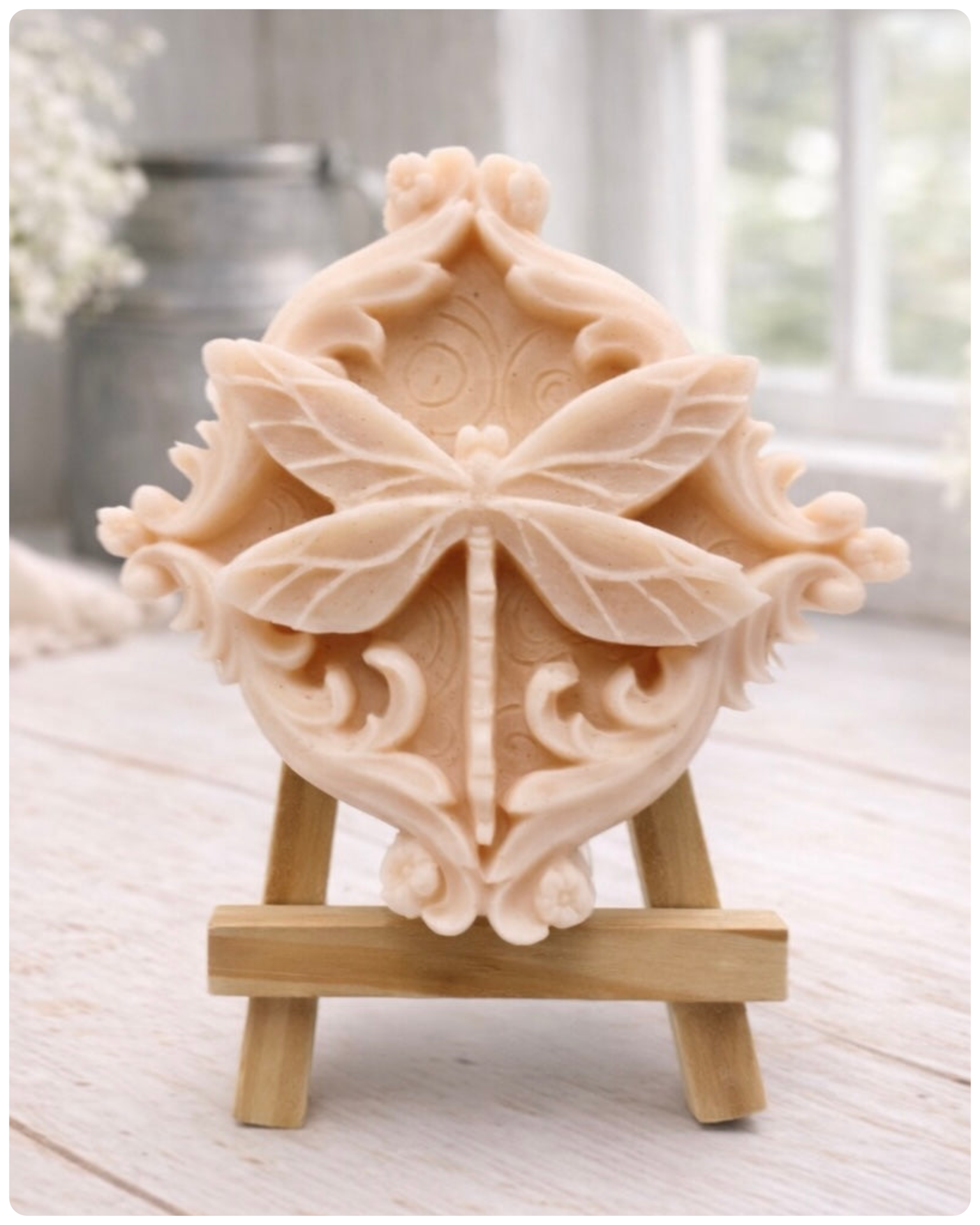 A beautiful handmade vegan soap in a natural tone, featuring a highly detailed relief of a dragonfly with delicate, outspread wings and a textured body, displayed on a miniature wooden easel against a soft farmhouse background.