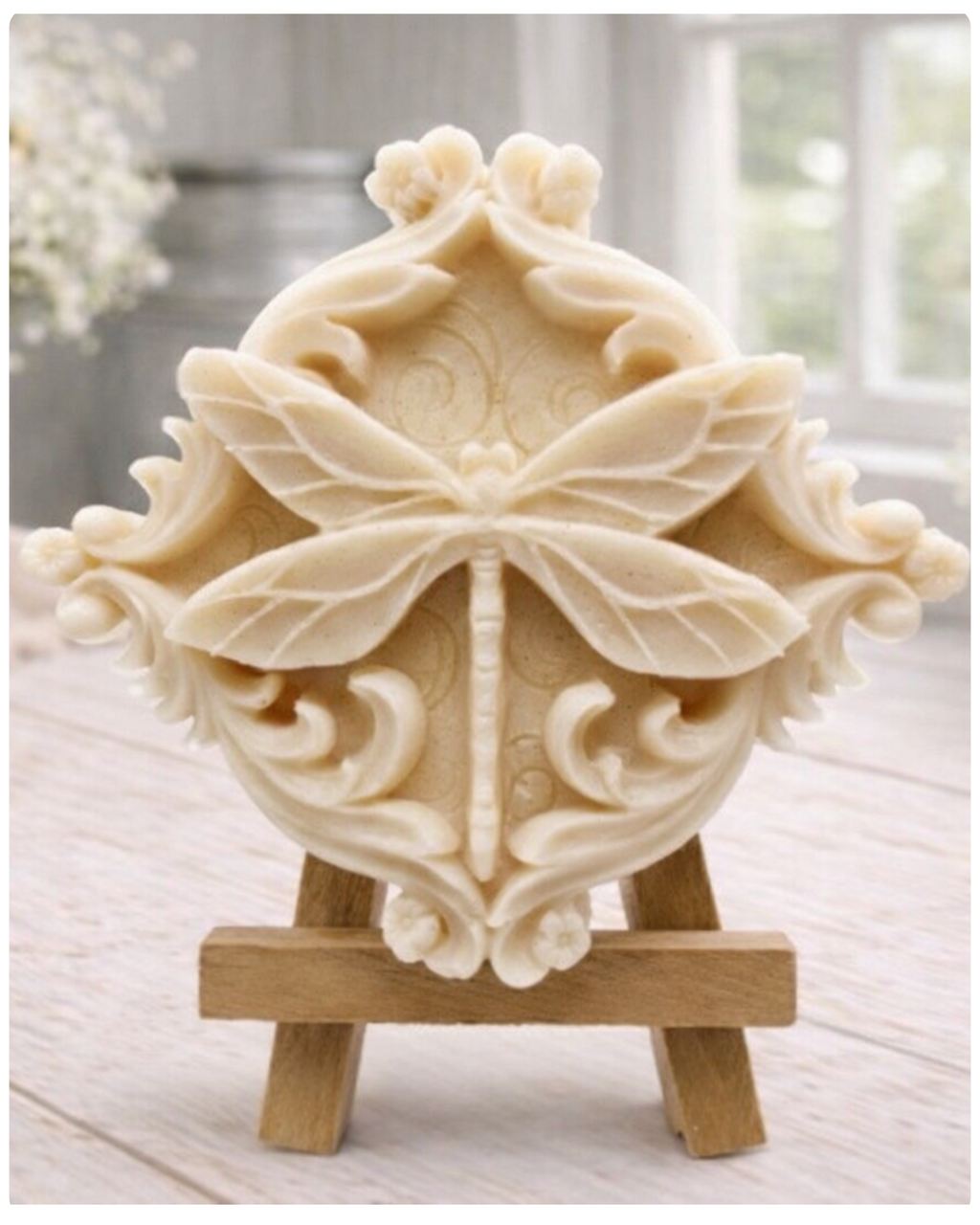 A beautiful handmade vegan soap in a natural tone, featuring a highly detailed relief of a dragonfly with delicate, outspread wings and a textured body, displayed on a miniature wooden easel against a soft farmhouse background.