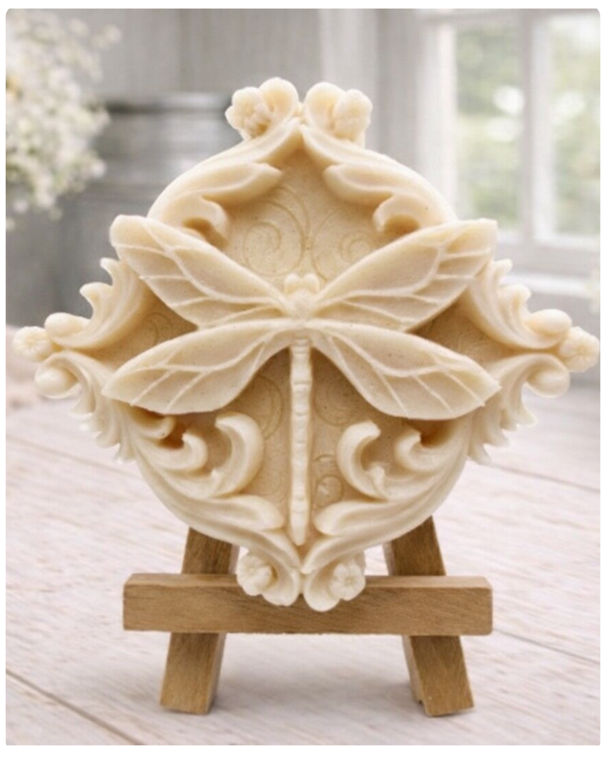 A beautiful handmade vegan soap in a natural tone, featuring a highly detailed relief of a dragonfly with delicate, outspread wings and a textured body, displayed on a miniature wooden easel against a soft farmhouse background.