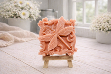 Load image into Gallery viewer, A beautiful handmade vegan soap in a natural tone, featuring a highly detailed relief of a dragonfly with delicate, outspread wings and a textured body, displayed on a miniature wooden easel against a soft farmhouse background.
