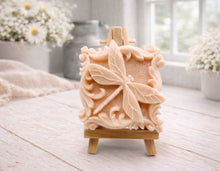 Load image into Gallery viewer, A beautiful handmade vegan soap in a natural tone, featuring a highly detailed relief of a dragonfly with delicate, outspread wings and a textured body, displayed on a miniature wooden easel against a soft farmhouse background.
