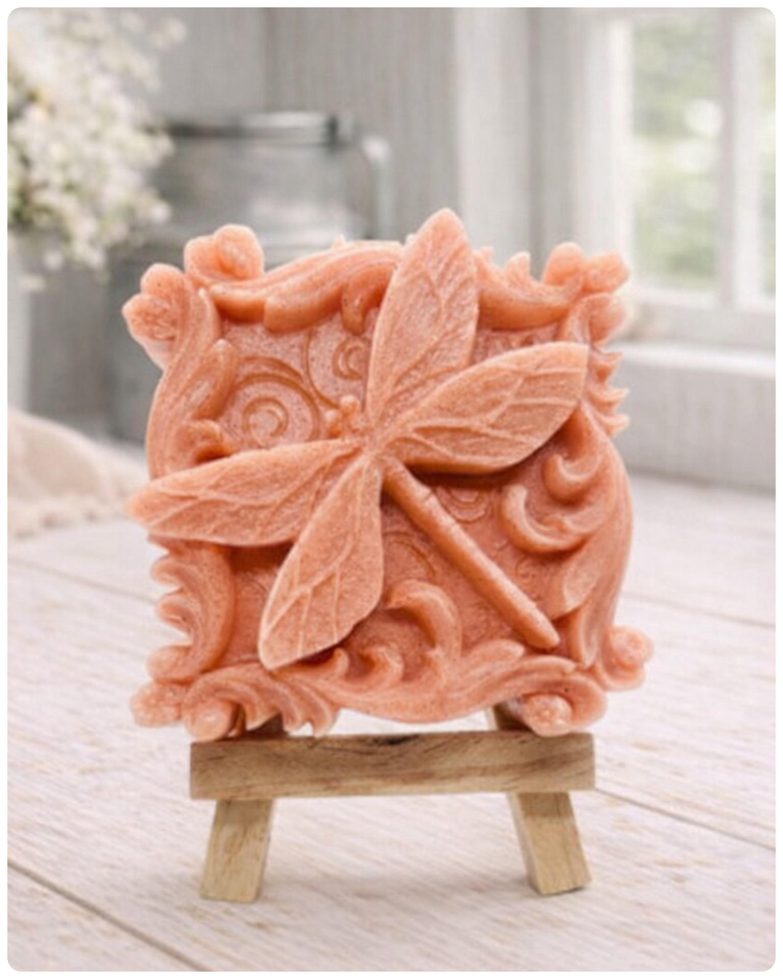 A beautiful handmade vegan soap in a natural tone, featuring a highly detailed relief of a dragonfly with delicate, outspread wings and a textured body, displayed on a miniature wooden easel against a soft farmhouse background.
