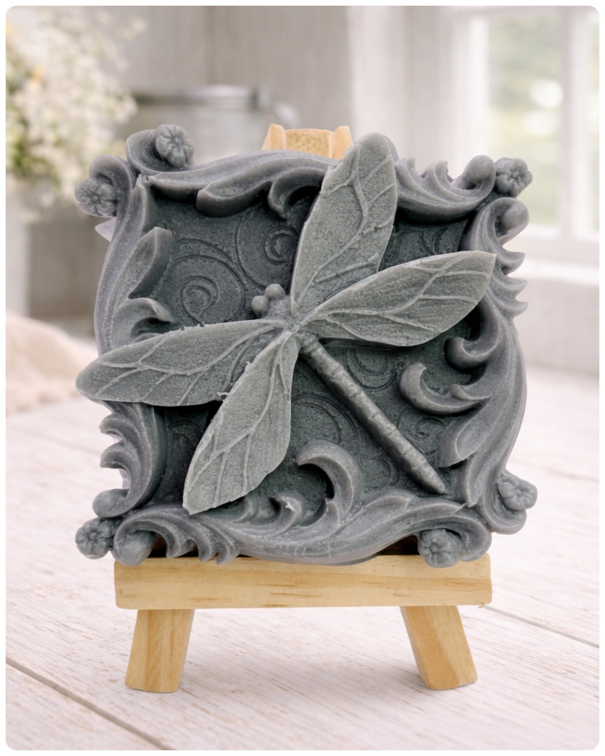 A beautiful handmade vegan soap in a natural tone, featuring a highly detailed relief of a dragonfly with delicate, outspread wings and a textured body, displayed on a miniature wooden easel against a soft farmhouse background.